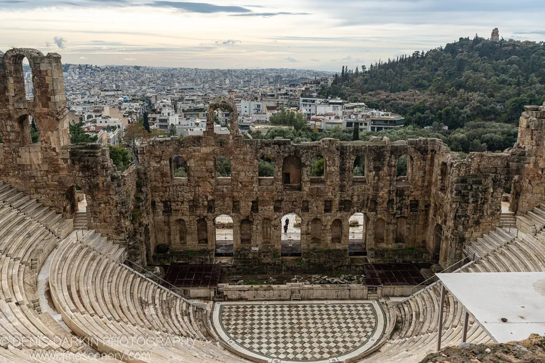 Marble seating tiers curve around a checkered stage floor at an ancient amphitheater above Athens under a winter sky.