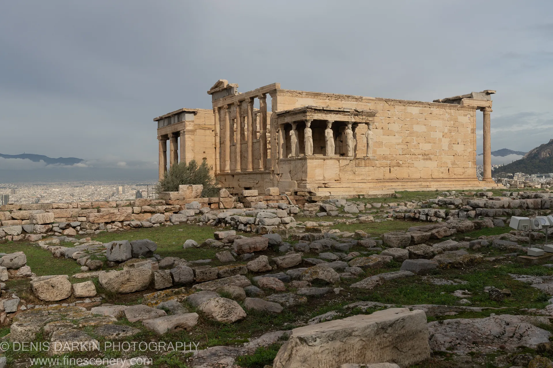 Weathered caryatid columns line the porch of an ancient temple, with Athens spread beyond under low winter cloud.
