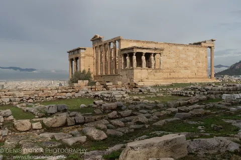 Weathered caryatid columns line the porch of an ancient temple, with Athens spread beyond under low winter cloud.