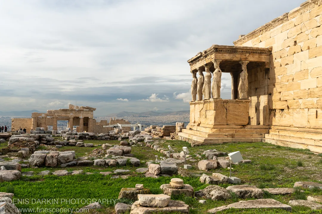 Ancient limestone ruins and a columned porch stand on a grassy plateau of the Acropolis beneath a soft overcast sky in Athens.