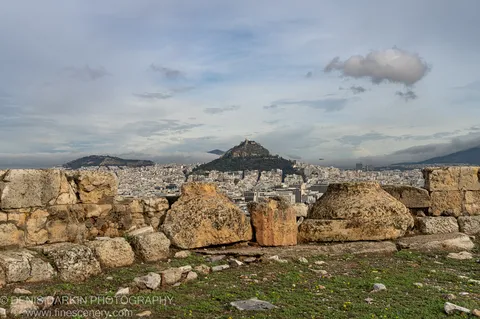 Weathered limestone fragments in the foreground give way to a dense Athens cityscape and a mountain peak beneath low cloud.
