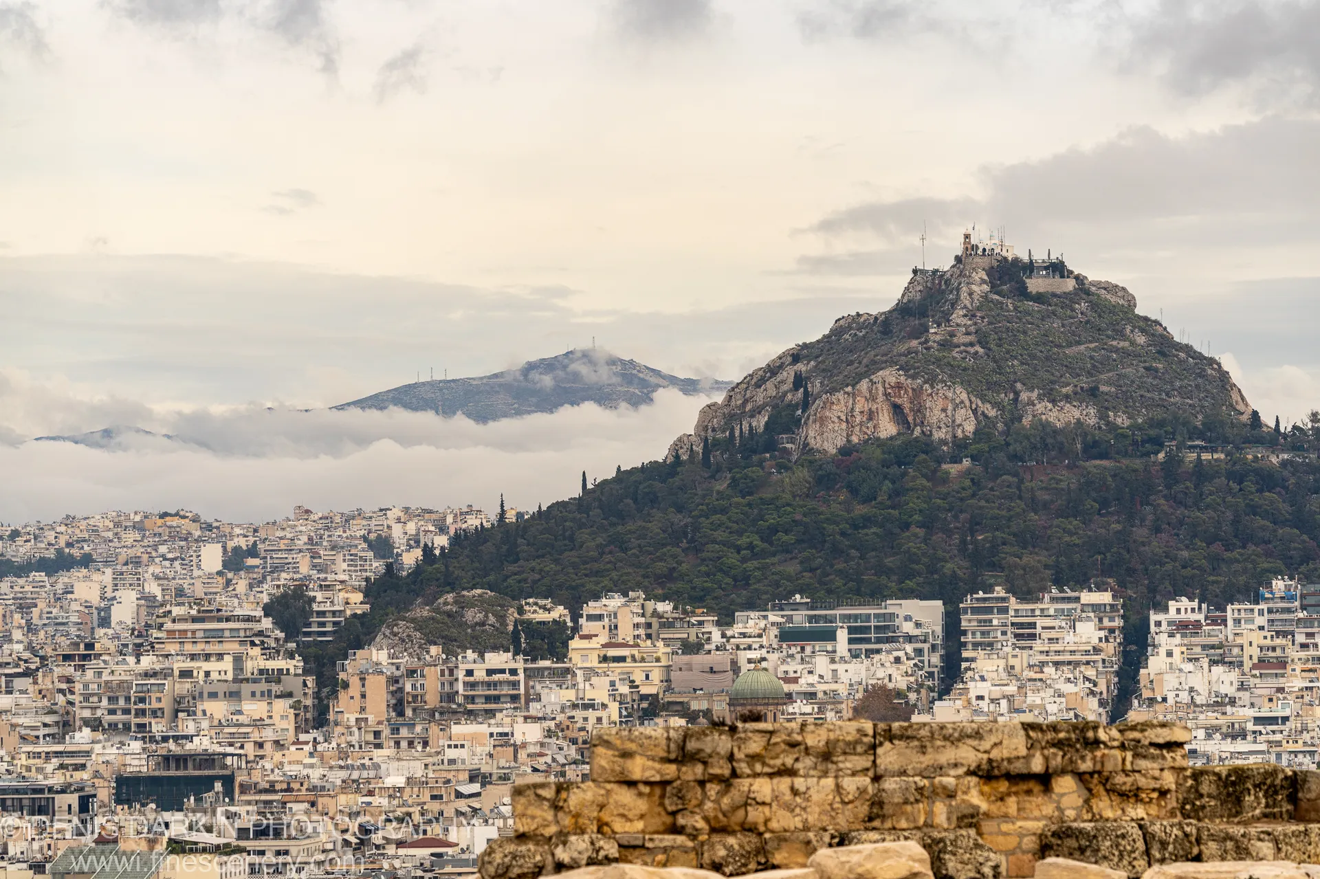 A textured ancient stone wall stands before the Athens cityscape, a verdant hillside with a historic structure at its summit in the distance.