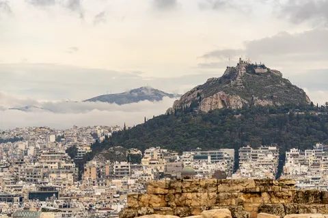 A textured ancient stone wall stands before the Athens cityscape, a verdant hillside with a historic structure at its summit in the distance.