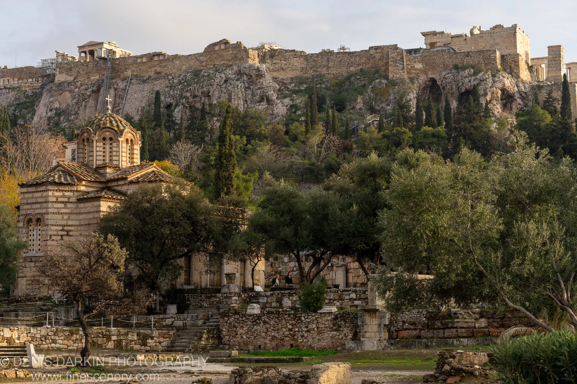 A stone church sits at the base of limestone cliffs topped with ancient ruins and cypress trees in Athens.