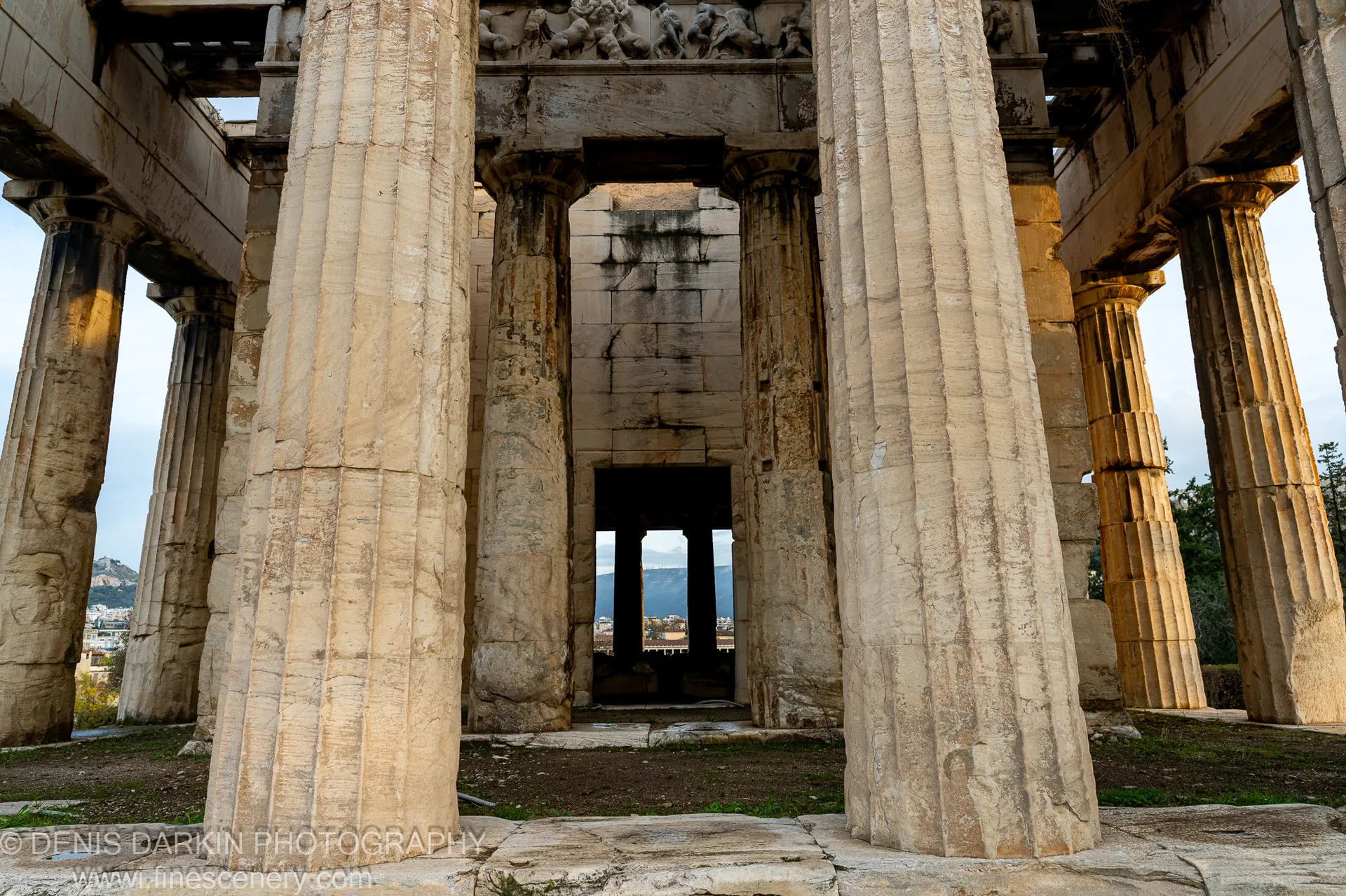 Massive fluted columns stand along an ancient temple portico in Athens, with a dark doorway opening toward distant hills.