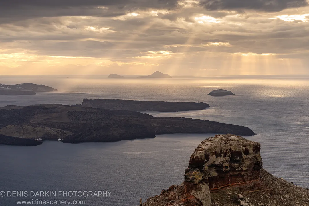 Rugged volcanic cliffs above the Santorini coastline look out over a sun-touched sea toward distant islands under a partly clouded sky.