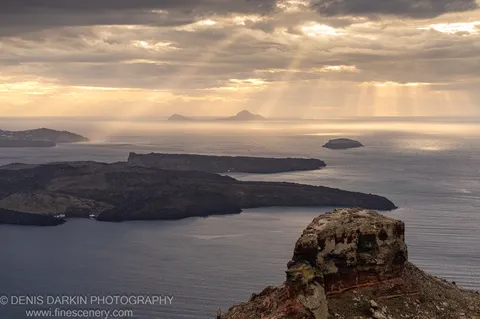 Rugged volcanic cliffs above the Santorini coastline look out over a sun-touched sea toward distant islands under a partly clouded sky.