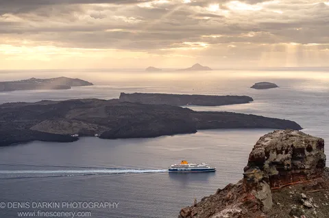 A rugged volcanic cliff edge at Santorini overlooks deep blue water where a ferry leaves a white wake below sunbeams breaking through cloud.