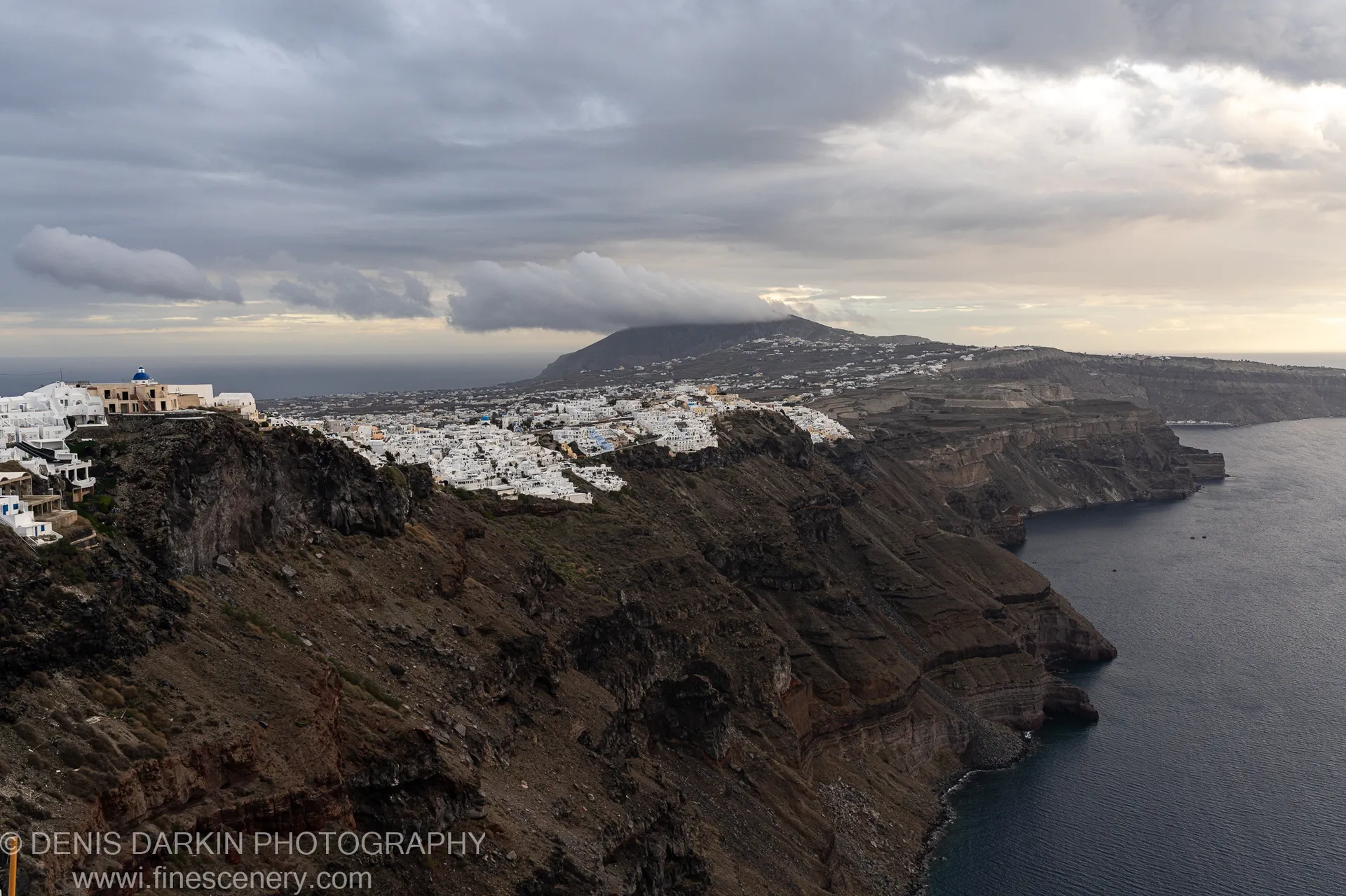Heavy clouds hang over barren volcanic slopes and the distant caldera sea, Santorini, December 2024.