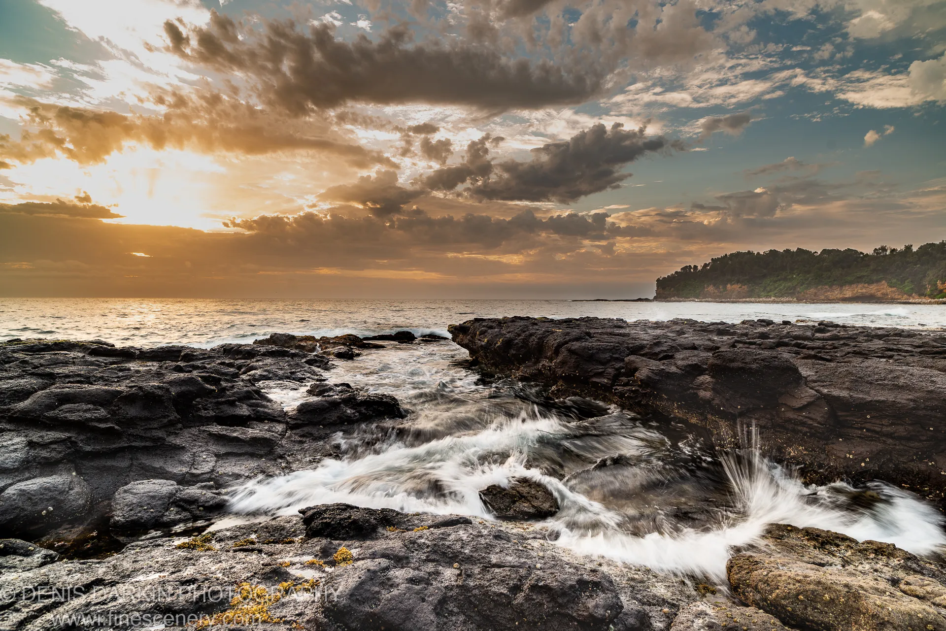 Golden dawn light breaks through heavy cloud above dark basalt rock as white water rushes through coastal crevices at Bendalong Point, New South Wales.