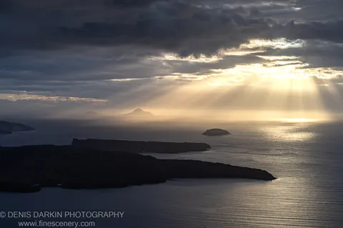 Sunbeams break through dark cloud over the caldera, casting warm light on the water and silhouetted headlands, Santorini. December 2024.