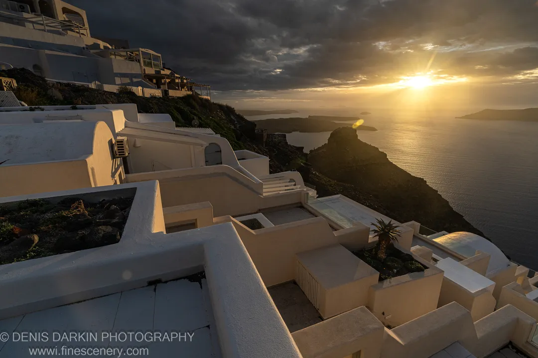 Shadowed white-washed buildings on a coastal cliff with a distant sunburst over dark water, Santorini, December 2024.