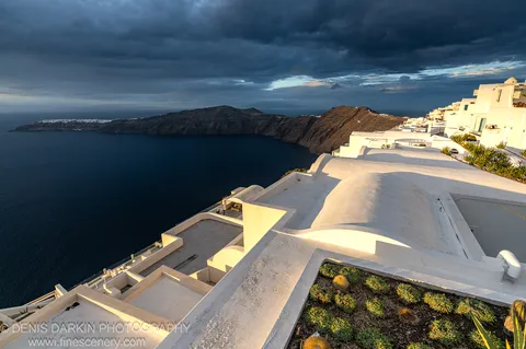 Sunlit white stucco rooftops and a succulent garden in the foreground, with dark volcanic caldera water and storm clouds beyond, Santorini, December 2024.