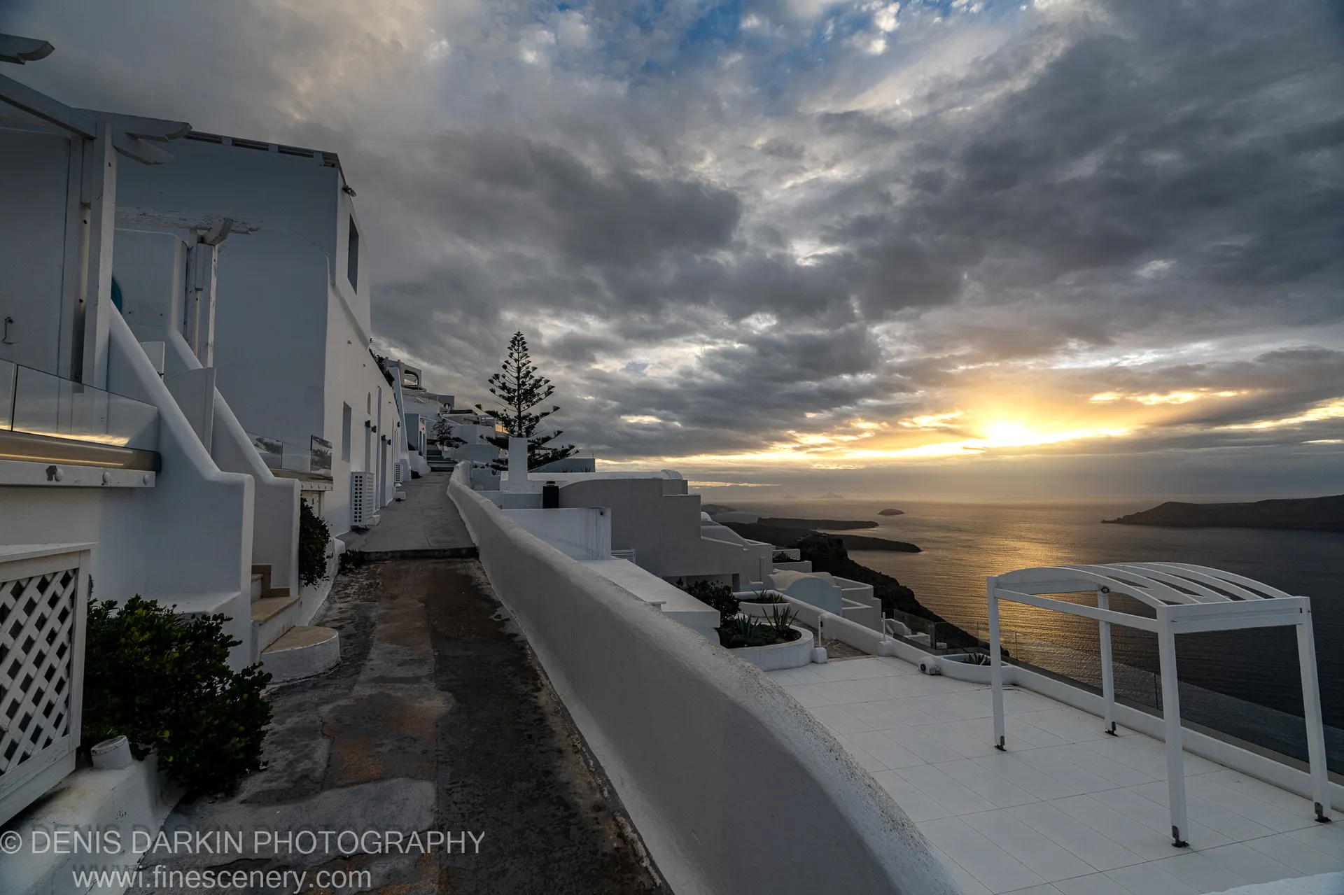 A coastal path leads down through white-washed terraced buildings toward a calm sea below heavy cloud, Santorini, December 2024.