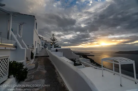 A coastal path leads down through white-washed terraced buildings toward a calm sea below heavy cloud, Santorini, December 2024.