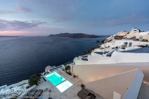 Lit swimming pools on a terraced cliffside villa above the Santorini caldera, with pastel dawn sky and a volcanic island on the horizon, December 2024.