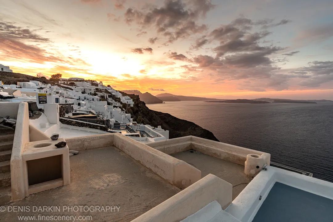 The dawn sky above Santorini shifts from deep blue to warm orange, silhouetting distant volcanic islands above the cliffside village, December 2024.