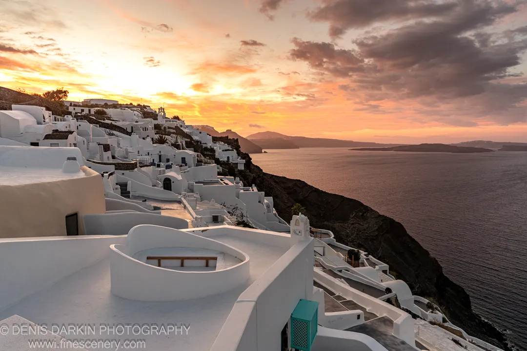 White-plastered terraces step down a dark volcanic cliffside in Santorini, warm sunrise light on the horizon beneath heavy winter clouds.