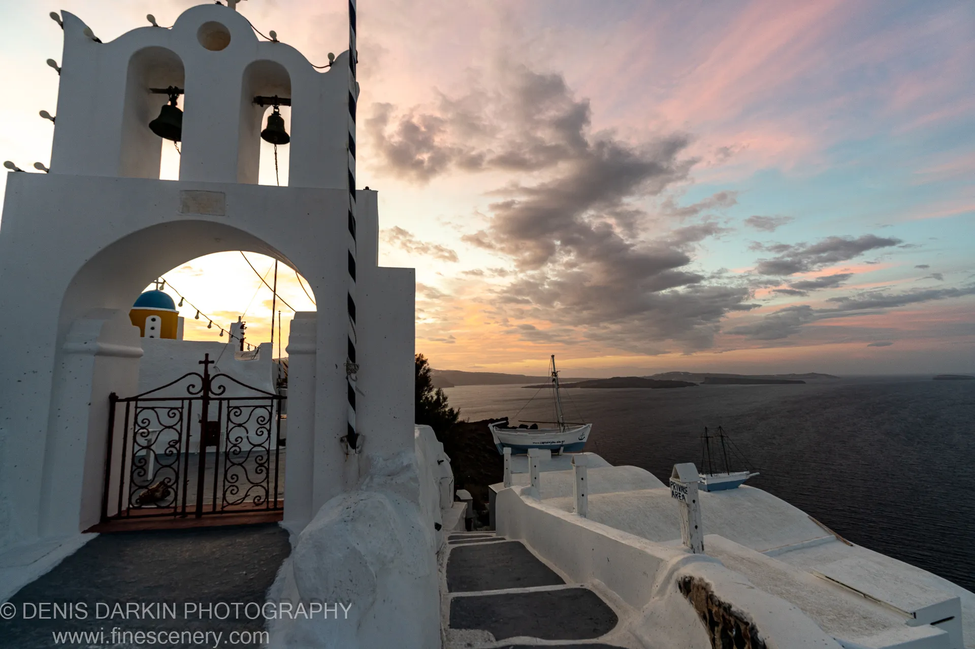 A white bell tower with an arched gateway stands in the foreground at Santorini, a blue dome visible against the pink-tinged sunrise sky behind.