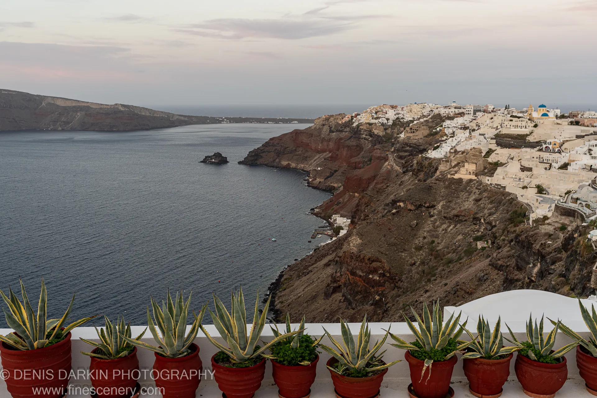 Terracotta pots of agave plants line a white ledge in the foreground, with white-washed buildings perched on dark volcanic cliffs above the Santorini caldera beyond.