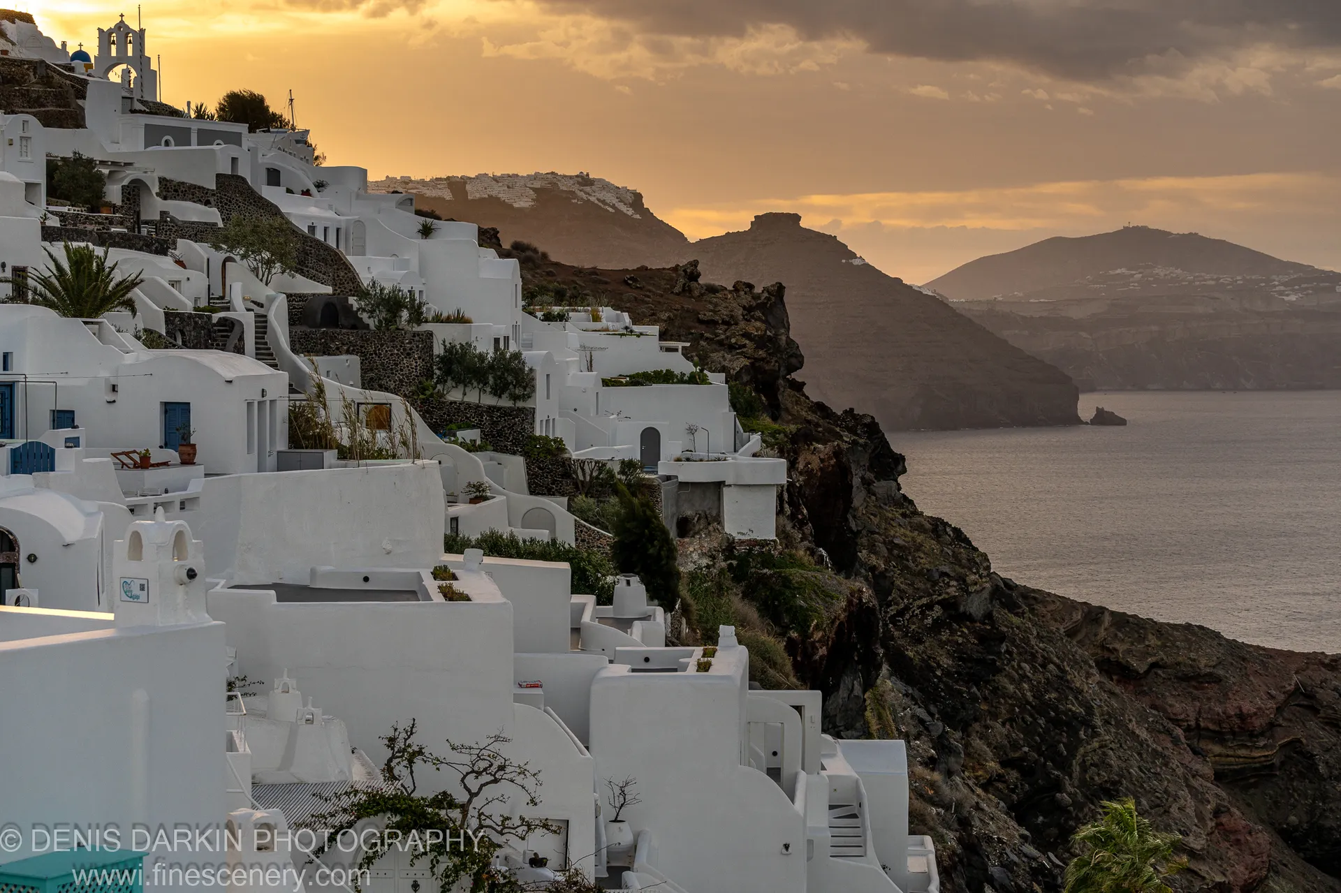 White-washed structures on a dark volcanic cliffside at Santorini, a church spire catching the warm sunrise light against a golden horizon.