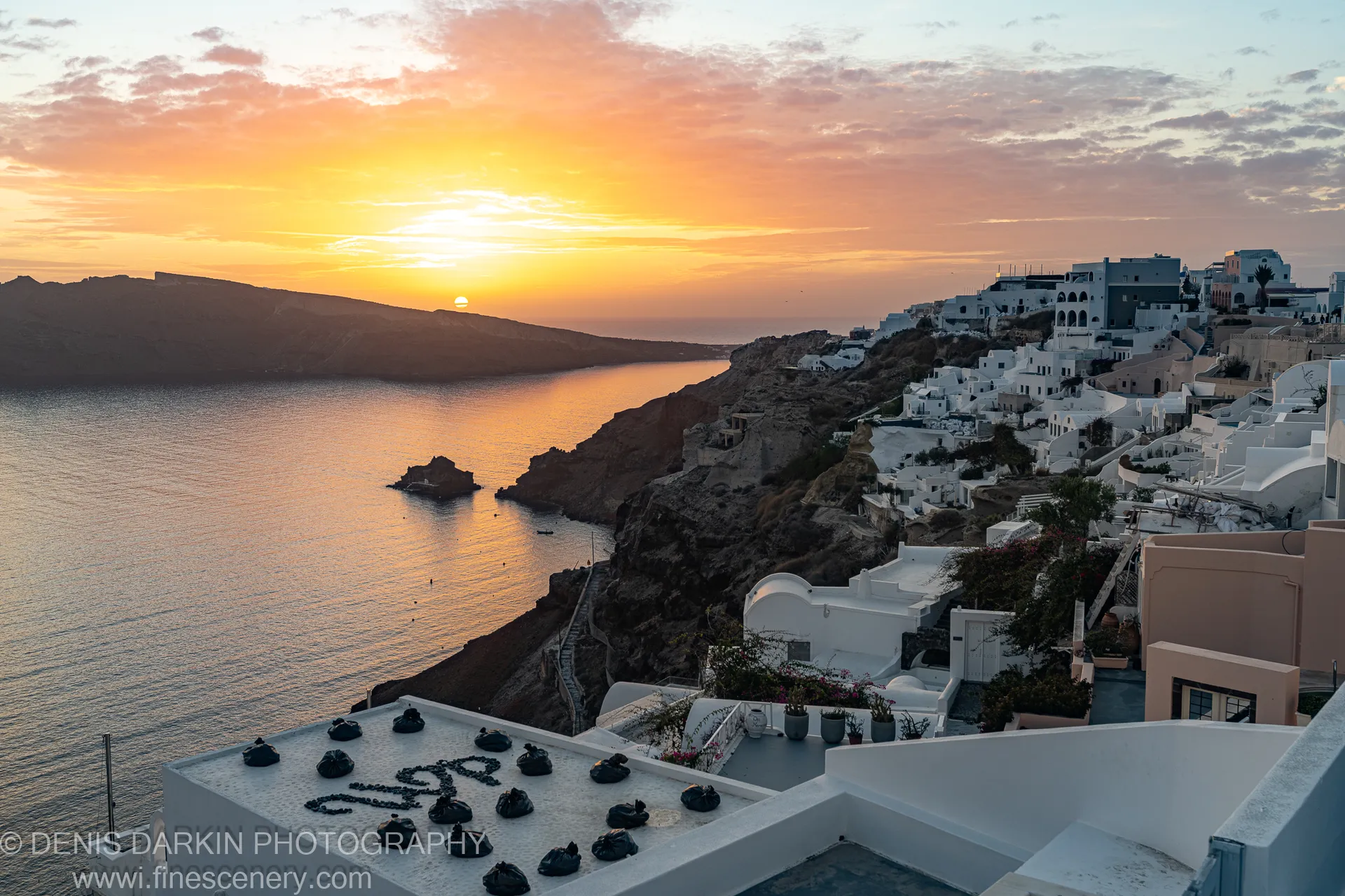 White cubic houses on a dark volcanic cliff at Santorini with warm afternoon light and an orange reflection on the calm caldera below.