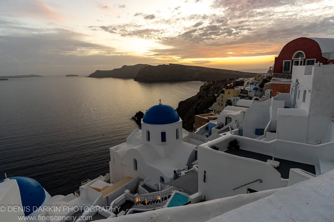 Vibrant blue and red domes on white plaster buildings at Santorini, with soft orange light at the horizon filtering through textured grey clouds at sunset.