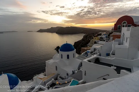 Vibrant blue and red domes on white plaster buildings at Santorini, with soft orange light at the horizon filtering through textured grey clouds at sunset.