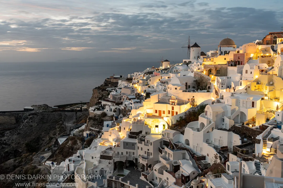 Traditional windmills silhouetted at sunset on the clifftop at Santorini, warm golden light from windows along the layered whitewashed village below and the sea beyond.