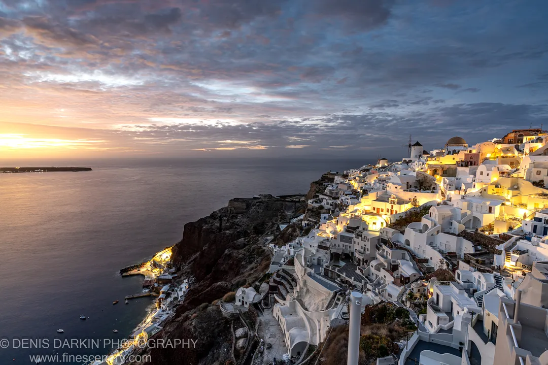 Warm lights from white-washed cubic structures on a dark cliffside at Santorini, the sky fading from orange to deep blue above the calm sea.
