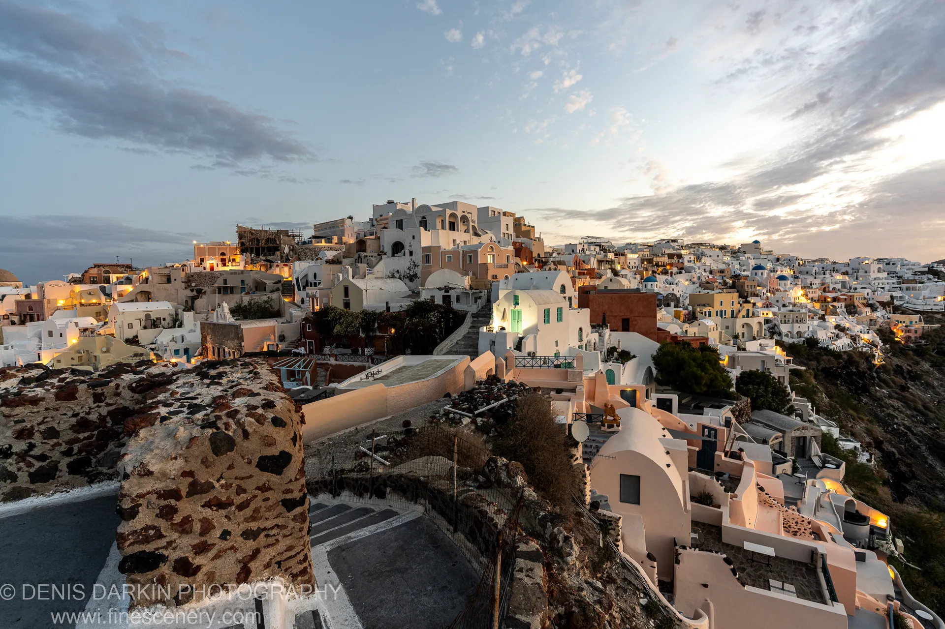 A hillside village at Santorini in the pre-dawn hour, warm amber lights from windows set against white-washed structures and a cool blue sky.