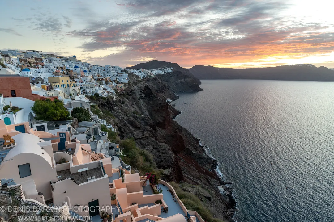 Whitewashed buildings with arched terraces step down a volcanic cliff face above the Santorini caldera at dawn, December 2024.