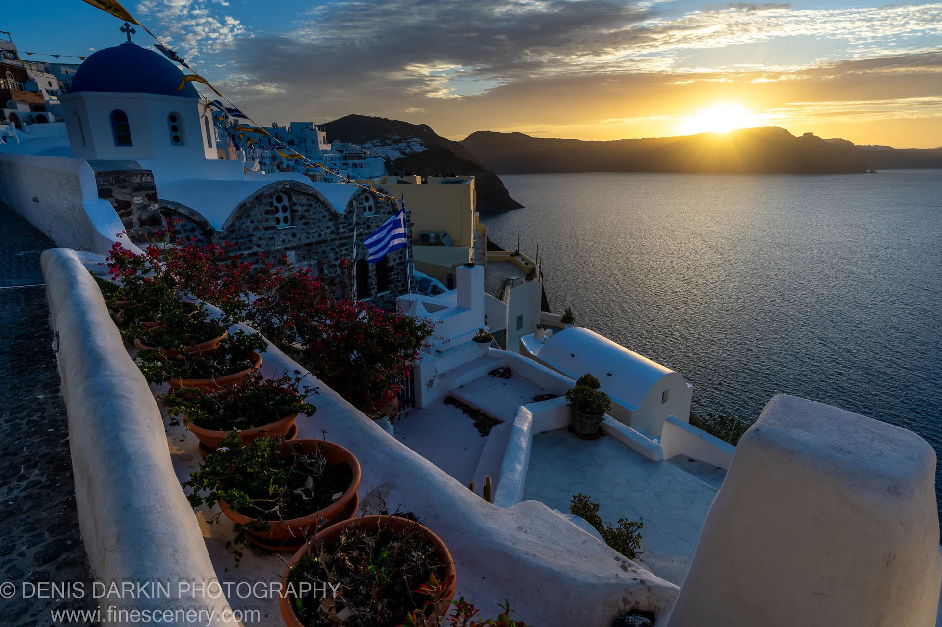 A blue-domed church with white plaster walls and fluttering flags stands above the caldera at dawn on Santorini, December 2024.