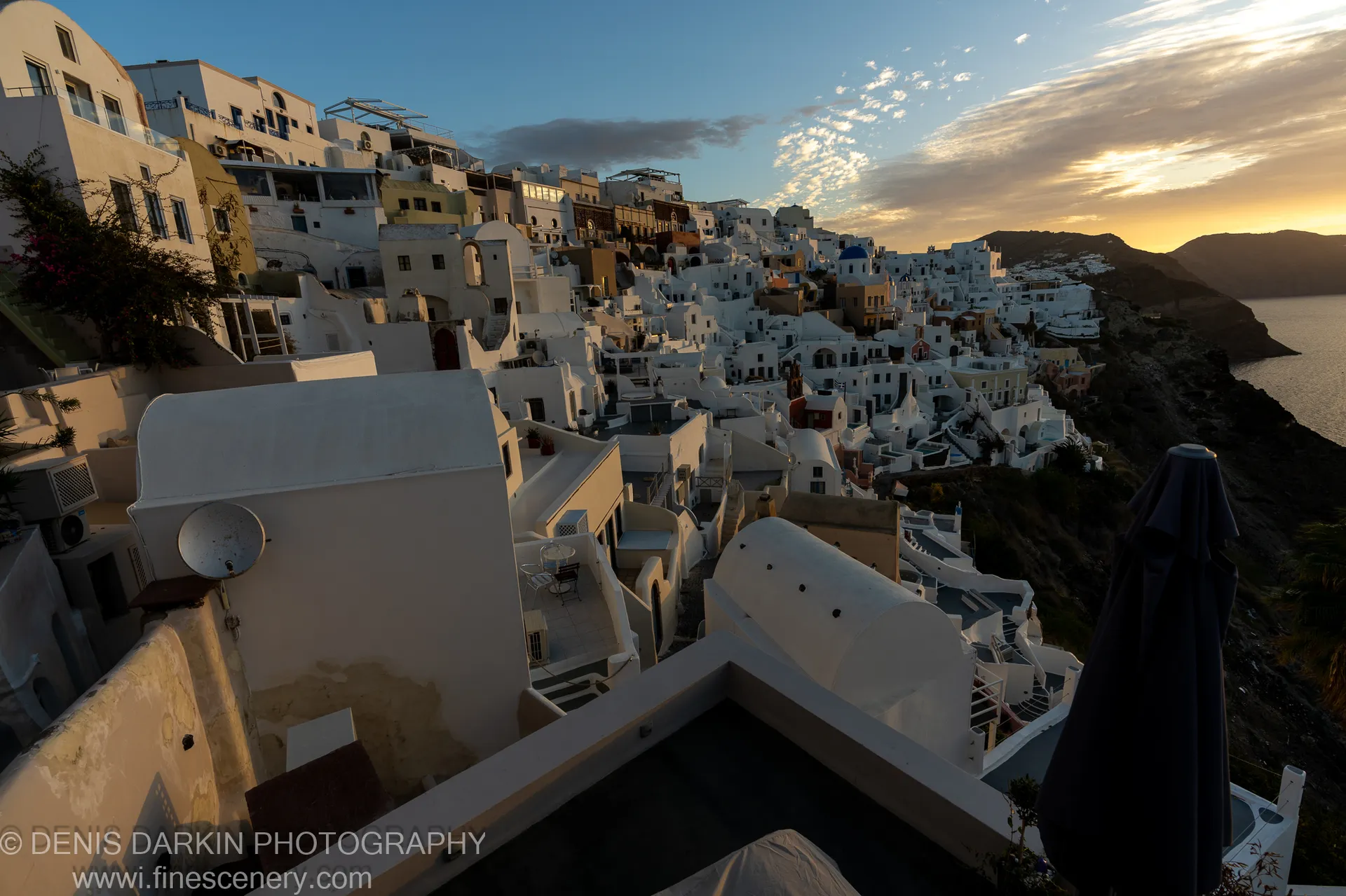 Wide-angle view of white cubic buildings stepping down the volcanic caldera rim at dawn in Santorini, December 2024.