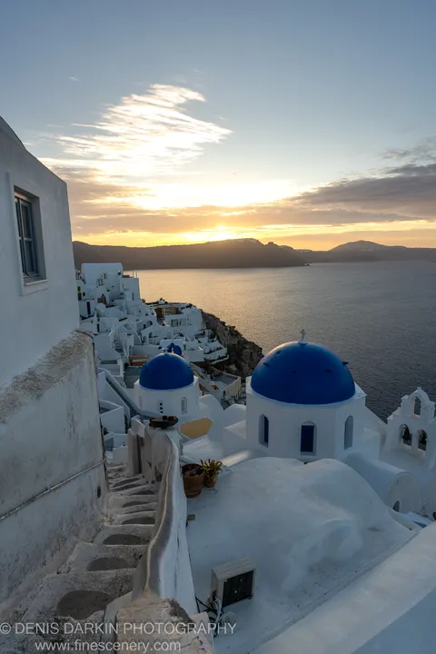 A weathered stone staircase descends toward the Santorini caldera at dawn, with cobalt-blue domes and orange-lit water below.