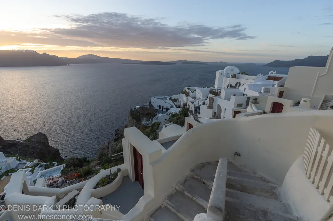 Whitewashed homes along a cliff edge at dawn in Santorini, winding stairs and curved walls in the foreground, December 2024.