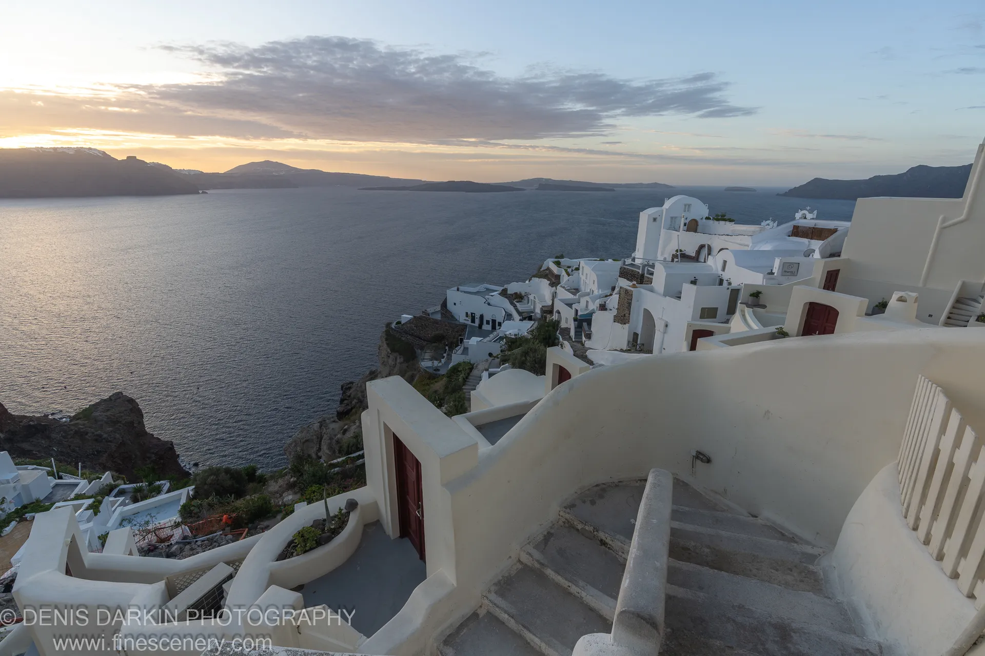 Whitewashed homes along a cliff edge at dawn in Santorini, winding stairs and curved walls in the foreground, December 2024.