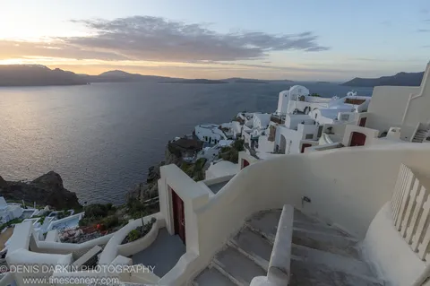 Whitewashed homes along a cliff edge at dawn in Santorini, winding stairs and curved walls in the foreground, December 2024.