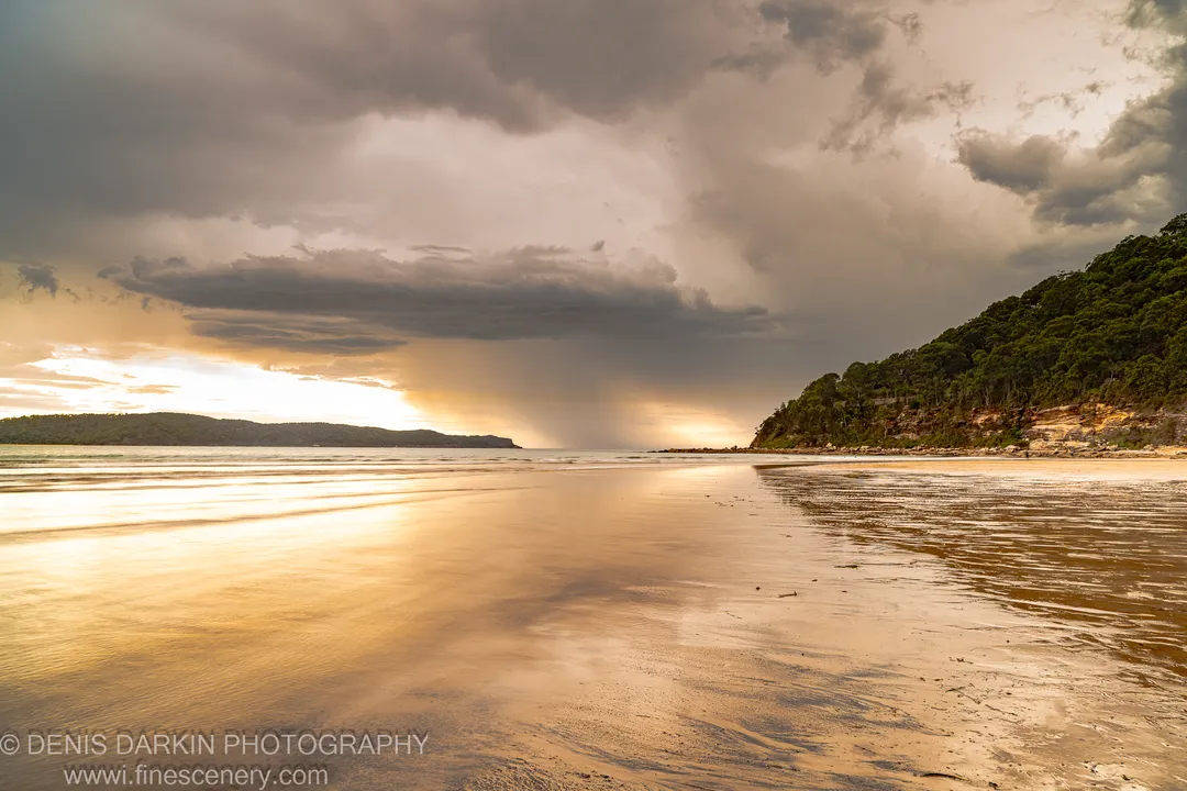 Rain shafts fall from dark storm clouds over the bay at Umina Beach, reflected in wet sand below a forested headland.