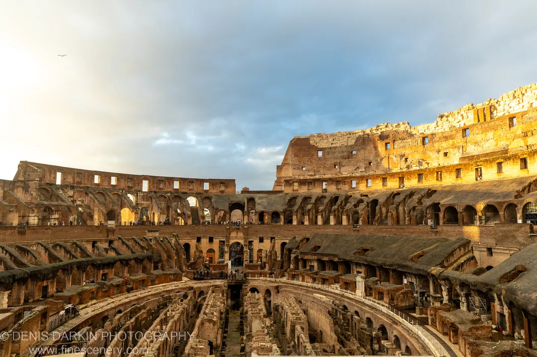 Sunlit upper walls of the Colosseum in Rome at golden hour, with the arena floor in deep shadow below, December 2024.