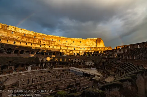 Warm sunlight on the Colosseum's upper stone facade, a faint rainbow in the dark storm sky above, Rome, December 2024.