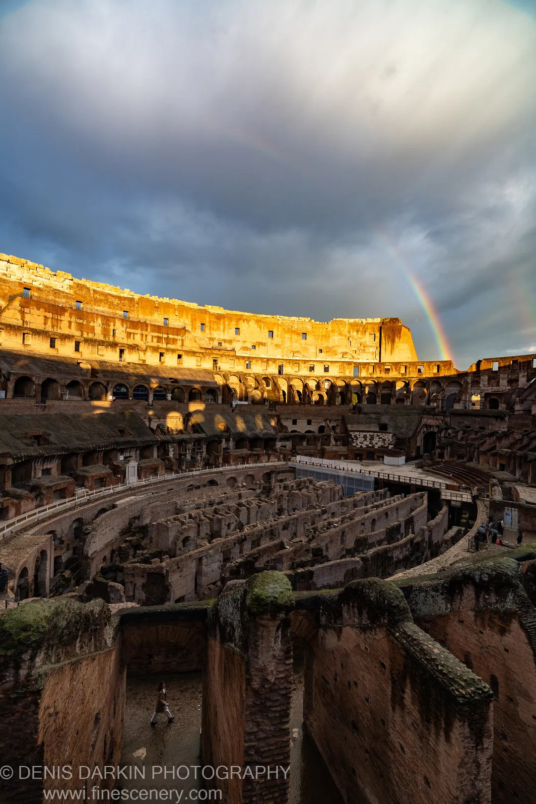 Inside the Colosseum in Rome; sunlit upper stone tiers above a shadowed arena floor, a winter rainbow in storm cloud above.