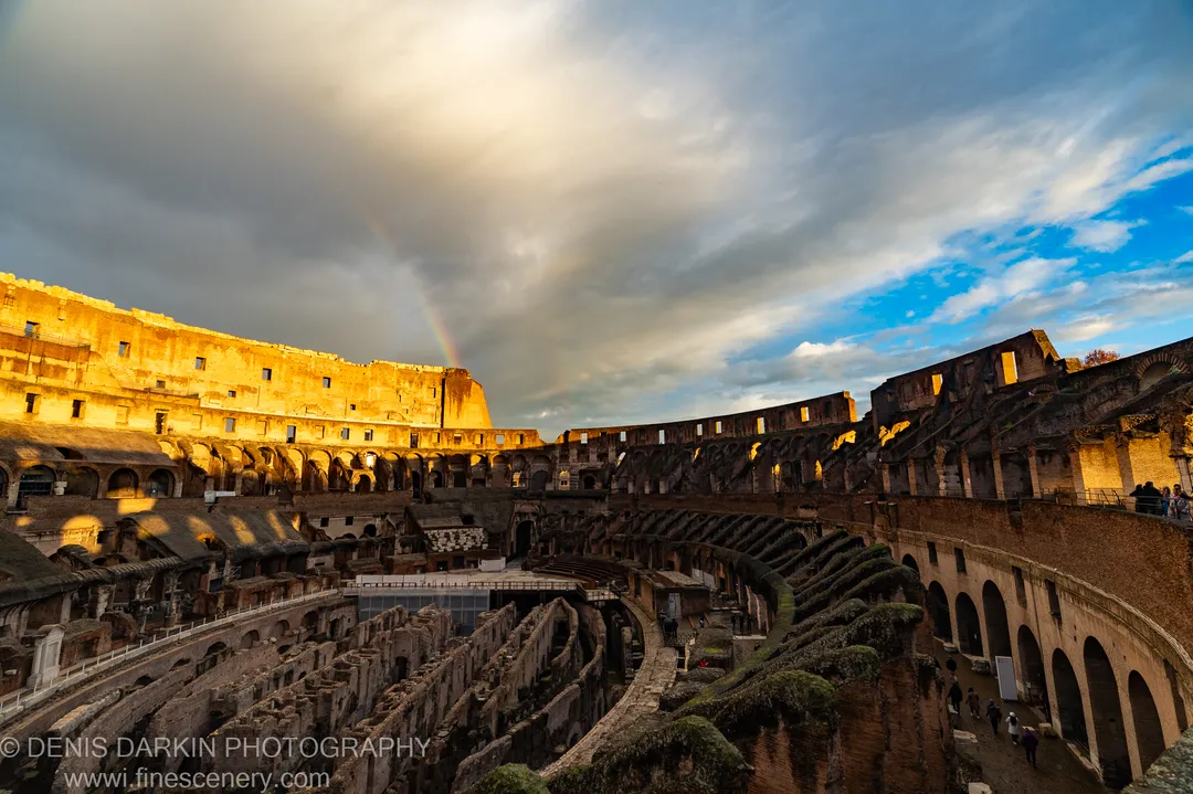 Upper stone wall of the Colosseum in Rome at winter golden hour, a rainbow arcing through storm cloud above the open arena.