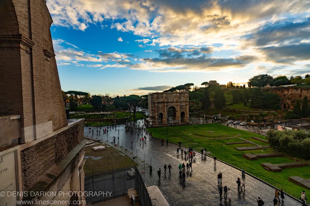Wet cobblestones and brick arches at golden hour in central Rome, small figures in the distance amid ruined masonry.