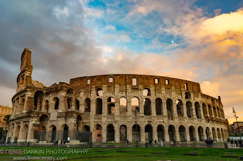 Colosseum exterior at golden hour in Rome, a pale moon in the sky above the curved arches, with cyclists and figures below.