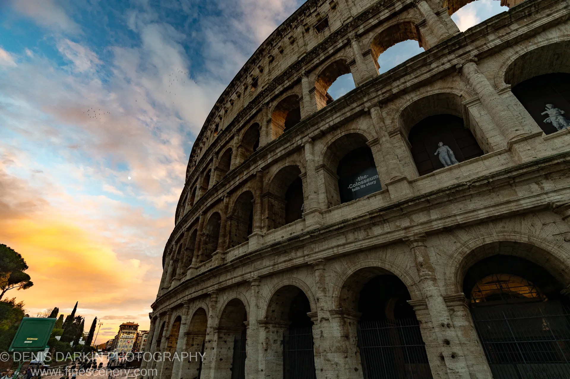 A flock of birds passes before the Colosseum's stone facade in Rome at golden hour, warm horizon glow on the ancient arches.