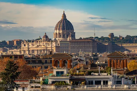 A domed basilica in Rome seen above terracotta rooftops in winter morning light, stone facade catching low sun against a clear sky.
