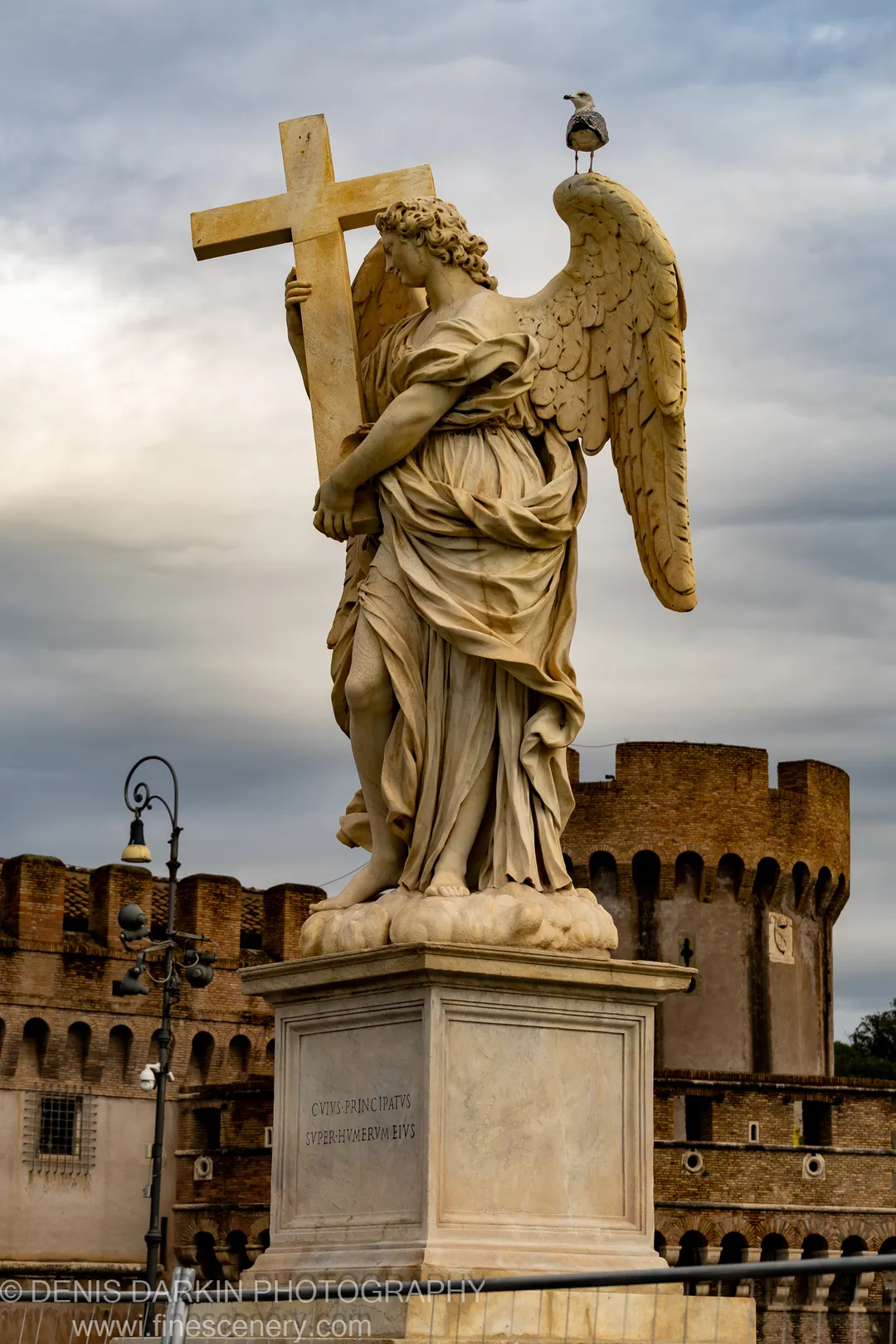 Marble angel with cross on the Ponte Sant'Angelo in Rome, a seagull perched on the wing's edge before the fortress walls.