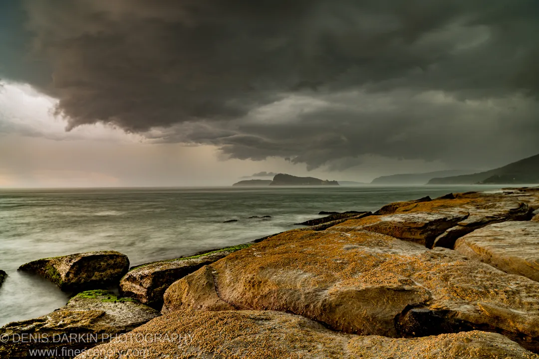 A shelf of flat, barnacle-covered sandstone rock at Umina Beach stretches toward a mist-shrouded horizon under a massive approaching storm cloud.