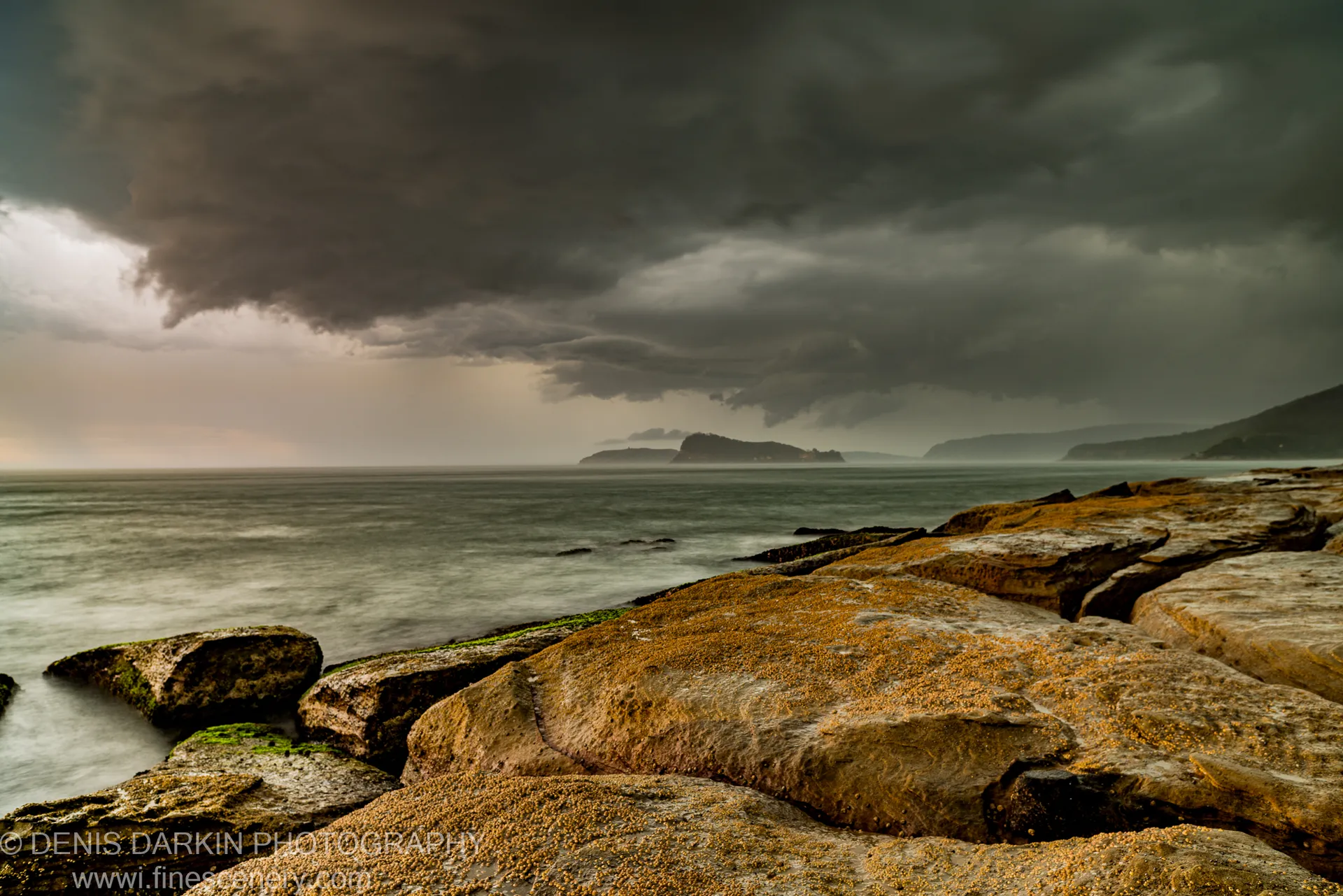A shelf of flat, barnacle-covered sandstone rock at Umina Beach stretches toward a mist-shrouded horizon under a massive approaching storm cloud.